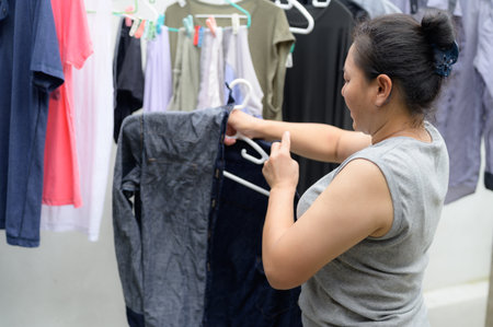 An adult Asian woman diligently performing her daily household routine, hanging clean clothes on hangers indoors. A candid moment reflecting home life and domestic activity.の写真素材