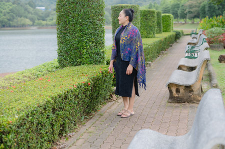 A serene mature Asian woman stands in a lush green park, enjoying the peaceful outdoor environment and natural daylight by the water.の写真素材