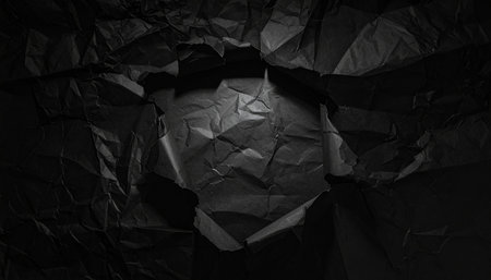 A dark, crumpled sheet of paper features a central torn hole, illuminated by a spotlight creating deep shadows.の素材