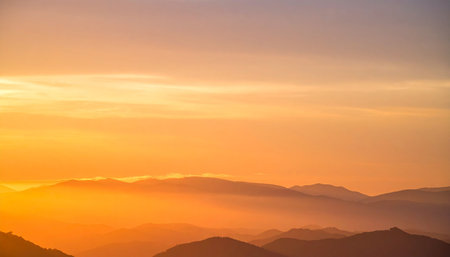 Distant showing distant mountain ridges shrouded in atmospheric haze at sunrise, with a warm orange glow...の素材