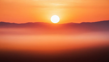 A bright sun descends behind a distant mountain range, with a glowing orange and yellow mist layer creating a...の素材