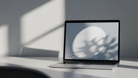 An open silver laptop rests on a white desk, with dramatic shadows cast by sunlight falling on the wall behind it.の素材
