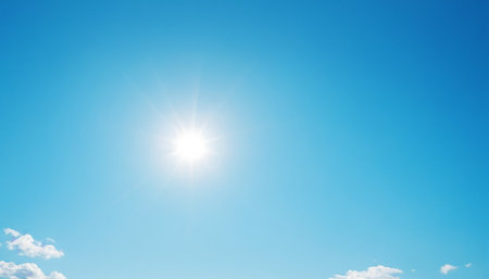 Lens showing sun with lens flare effect shines in a clear, vibrant blue sky with a few small white clouds in the...の素材