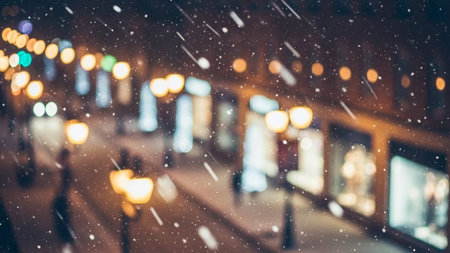 A blurry nighttime city street scene with falling snow and warm bokeh lights from streetlamps and shop windows...の素材