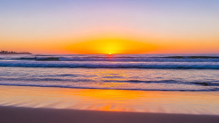 A tranquil beach scene at sunset with gentle waves washing onto the shore, reflecting the warm orange sky.の素材