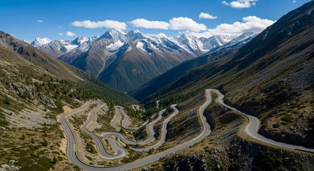An aerial view of a dramatic alpine landscape featuring a winding mountain pass road that snakes its way through steep, rugged slopes. The road is characterized by numerous curves and switchbacks, creating a serpentine pattern as it ascends through the valley. snow-capped mountains rise in the background under a clear blue sky. The surrounding slopes are a mix of rocky terrain and sparse vegetation, highlighting the challenging and nature of this high-altitude environment.の素材
