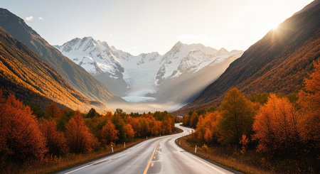 A winding asphalt road carves its way through a mountain valley during the peak of autumn. Towering, snow-capped mountains and a distant glacier frame the scene, while the slopes are ablaze with the vibrant colors of fall foliage in shades of orange, red, and yellow. Sunlight streams through the sky, casting dramatic rays and illuminating the mist in the valley.の素材