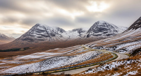 A winding asphalt road with a metal guardrail carves its way through a rugged mountain landscape in the Scottish Highlands. The peaks of the mountains are dusted with snow, contrasting with the dry, brown and orange hues of the heather and grass in the valley. The sky is overcast and cloudy, adding to the dramatic and moody atmosphere of the scene. The captures the raw beauty and isolation of the Scottish wilderness.の素材