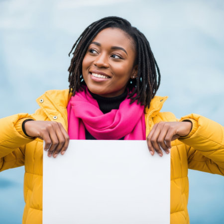 A cheerful young Black woman with dreadlocks is smiling and looking to the side while holding a blank white sign. She is wearing a bright yellow jacket and a vibrant pink scarf, isolated on a white background.の素材