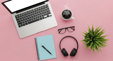 A flat lay of a laptop, notebook, pen, coffee cup, headphones, glasses, and a small plant on a pink surface.の素材