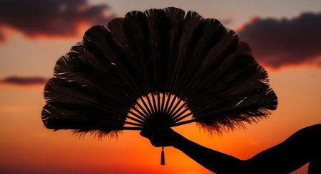 A dark silhouette of a person's arm and hand holding a large, ornate feather fan. The fan is spread open against a vibrant sunset sky.の素材