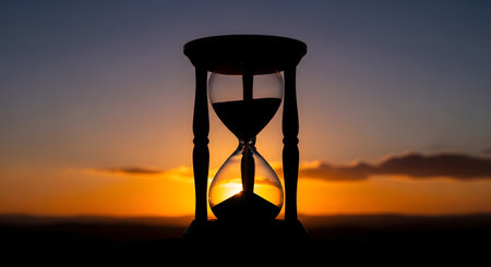 A dark hourglass stands in silhouette against a fiery orange and purple sunset, symbolizing the passage of time.の素材