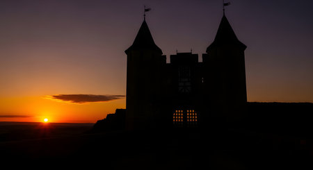 A dramatic silhouette of a castle with twin spires stands against a fiery orange and purple sunset, casting a mystical glow.の素材