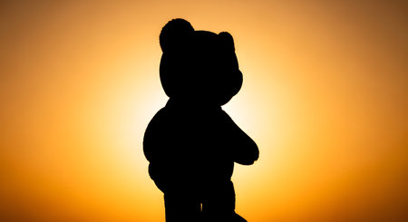 A stark black silhouette of a teddy bear stands against a vibrant, warm orange gradient background, creating a striking contrast.の素材