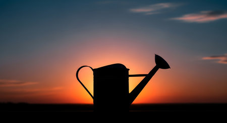 A dark silhouette of a classic watering can stands against a dramatic sunset, casting a warm glow.の素材