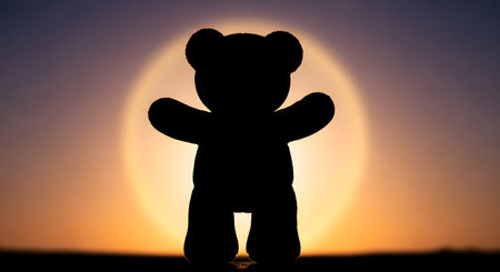 A dark silhouette of a teddy bear stands against a glowing, warm sunset, its arms raised in a welcoming gesture.の素材