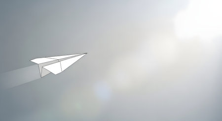 A simple white paper airplane flies gracefully across a vast, sunlit sky with soft clouds and lens flare.の素材