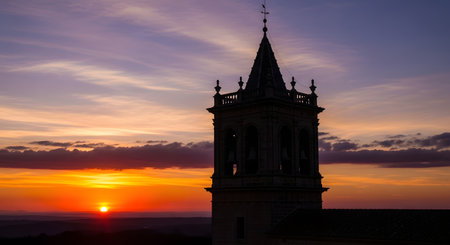 A tall, gothic tower stands in silhouette against a vibrant sunset with orange, yellow, and purple hues painting the sky.の素材