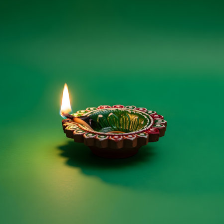 A single, ornate diya burns brightly with a warm flame, casting a soft glow on a plain green surface.の素材