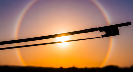 A violin bow is silhouetted against a vibrant sunset, with a sun halo creating a celestial spectacle in the sky.の素材