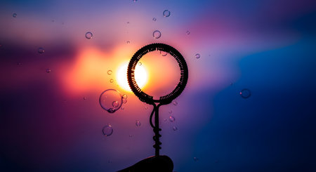 A close-up shot of a bubble wand creating iridescent bubbles against a vibrant sunset sky with streaks of pink, orange, and blue.の素材