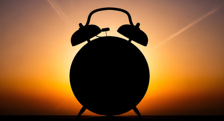 A stark black silhouette of a vintage alarm clock stands against a vibrant orange and yellow sunset, casting long shadows.の素材