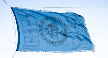A light blue flag with a detailed emblem and fringed edges flutters against a clear, bright sky.の素材