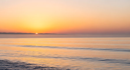 A serene sunrise paints the sky in warm hues of orange and yellow, casting a soft glow on the tranquil ocean and its gentle waves.の素材