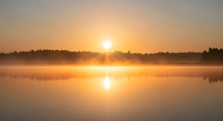 A radiant sun breaks through morning mist, casting a warm golden glow over a tranquil lake and silhouetted forest.の素材