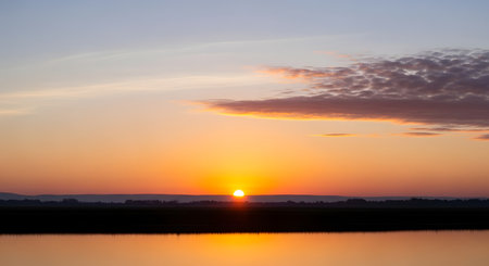 The sun dips below the horizon, casting a warm orange glow across the sky and reflecting on the tranquil water.の素材