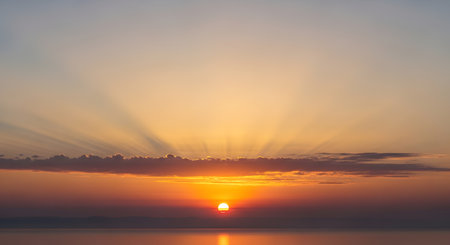 The sun dips below the horizon, casting warm golden rays through scattered clouds over a tranquil sea.の素材