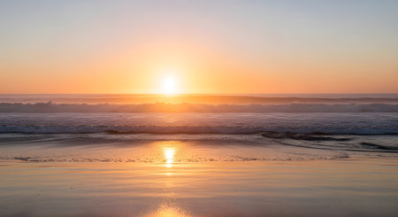 A vibrant sunset casts a warm golden glow across the ocean and wet beach sand, creating a serene and beautiful natural scene.の素材