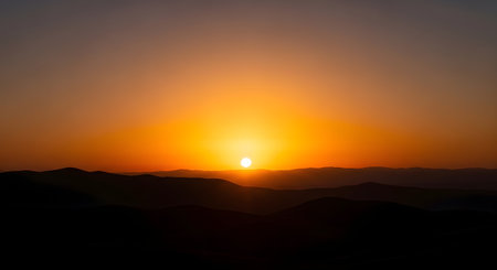 A vibrant orange sun dips below a dark, silhouetted mountain range, casting a warm glow across the sky.の素材