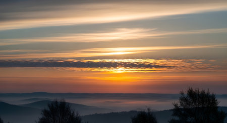 A vibrant sunrise paints the sky with warm colors, casting light over misty mountains and dark, silhouetted trees.の素材