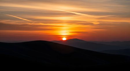 The sun sets behind silhouetted mountains, casting a vibrant orange glow across the sky with wispy clouds.の素材