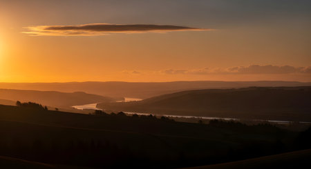 A breathtaking sunset casts a warm golden glow over silhouetted rolling hills and a winding river below.の素材