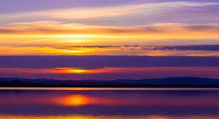 A breathtaking sunset paints the sky with vibrant oranges, yellows, and purples, reflecting on the tranquil water below. Distant mountains are silhouetted against the colorful horizon.の素材