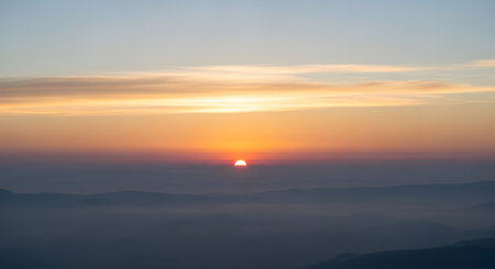 The sun rises with a brilliant orange glow, casting warm light over a vast expanse of misty, blue-tinged mountains.の素材