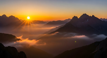 Dramatic mountain range silhouette against a vibrant sunrise, with golden light illuminating swirling clouds and mist below.の素材