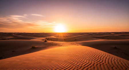 Vast desert landscape with rippling sand dunes bathed in the warm glow of a setting sun and a colorful, cloudy sky.の素材