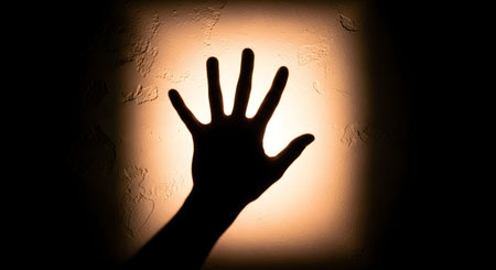 A dark silhouette of an open hand with fingers spread is pressed against a textured, illuminated surface, creating a dramatic and mysterious effect.の素材