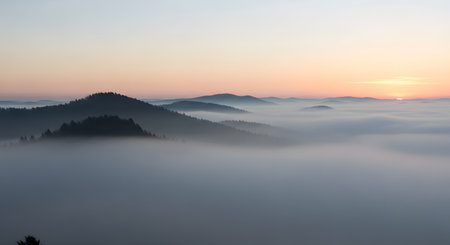Silhouetted mountain peaks emerge from a thick blanket of fog as the sun rises, casting a soft orange glow across the sky.の素材