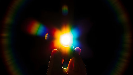 A person is holding a prism in their hands, creating a rainbow effect in the dark. The prism is refracting light, producing a colorful display around it.の素材
