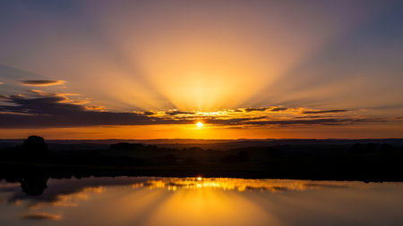 A serene sunset scene with the sun's rays bursting through the clouds, casting a warm glow over a tranquil lake. The sky is painted with hues of orange and yellow, reflected perfectly on the calm water's surface, creating a mirror-like effect. Silhouettes of trees and hills line the horizon, adding depth to the picturesque landscape.の素材