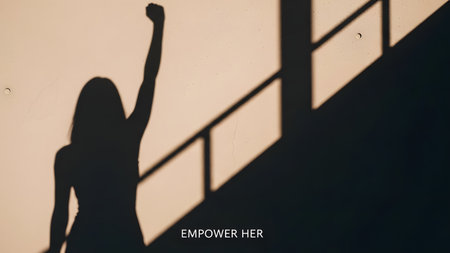 A silhouette of a woman standing with her right arm raised in a fist, near a staircase, conveying a sense of empowerment and strength. The image is in a warm, sepia tone, with the text 'EMPOWER HER' at the bottom.の素材