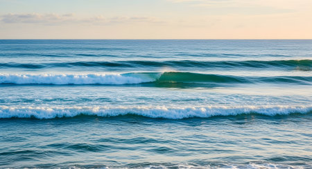 A serene ocean scene with waves gently rolling towards the shore, creating a soothing atmosphere. The water is a mix of deep blue and turquoise hues, with white foam on the surface. The sky above is a soft blend of blue and pale yellow, indicating a peaceful time of day, possibly during sunrise or sunset.の素材