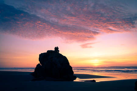Two silhouetted people sit on rock by seashore and watch a glorious sunset.の写真素材