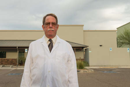 Aged physician in white lab coat outdoors in front of clinic.の写真素材