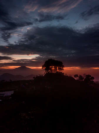 Silhouette of Trees growing on the top of a small hill against the background of the sky at sunrise with cloudy weather. Silancur Highland, Magelang, Indonesiaの写真素材