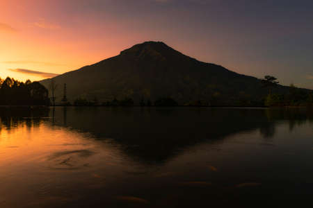 Sunrise with Mount Sumbing with lake surface on the foreground. The lake surface make reflection of mountain and sunrise sky. Embung Kledung, Central Java, Indonesiaの写真素材